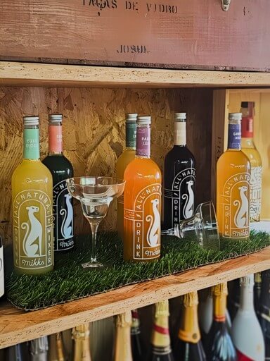 Várias garrafas Mikks Drinks com dois copos de margarita sobre uma estante de cortiça da garrafeira Black Pepper and Basil, com tapete de relva artificial por baixo